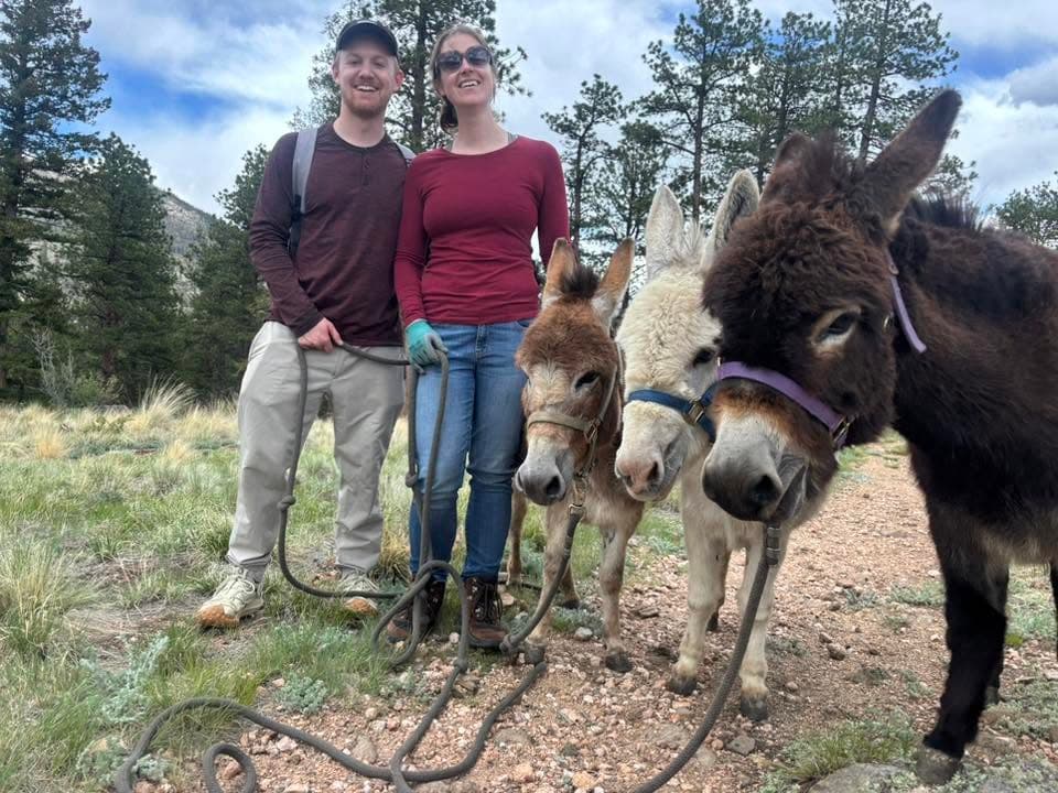 Two hikers smiling with three miniature donkeys on a mountain trail near Bailey, Colorado